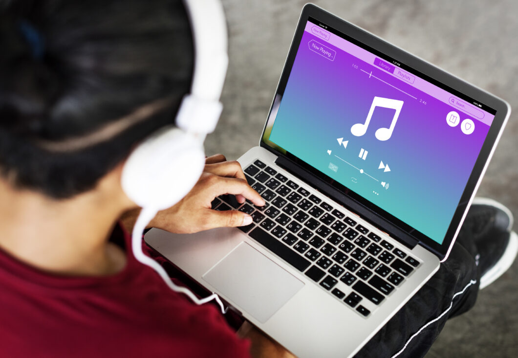 Foto de uma pessoa utilizando um notebook, usando um fone branco, com uma ilustração de um app de música na tela do aparelho