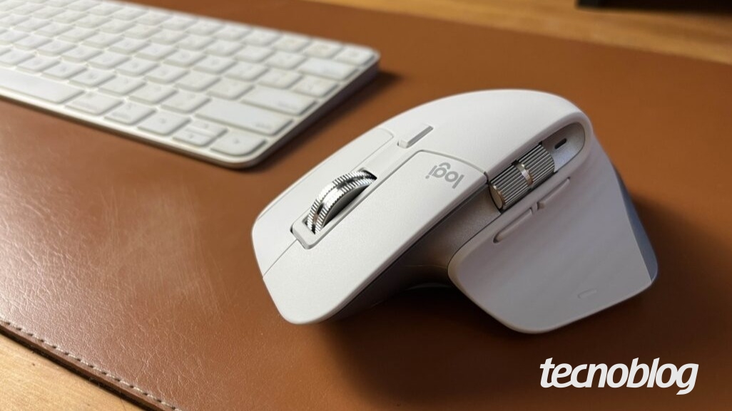 Imagem do mouse MX Master 3S da Logitech de cor branca, em cima de uma superfície de couro marrom. O mouse é posicionado de forma oblíqua na superfície, com a parte inferior à vista. A sua cor é branca, com detalhes em prata nos botões e roletes laterais. A marca "Logi" está visível na parte superior do rato. Um teclado branco também é visível no canto superior esquerdo da imagem, fora de foco. A superfície é de couro sintético marrom, com textura visível. Na parte inferior direita, está o logo do Tecnoblog.