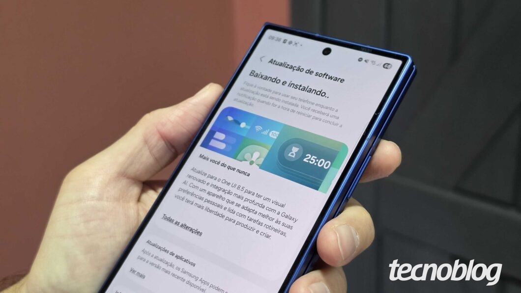 Uma mão segura um smartphone com bordas azuis metálicas, exibindo a tela de "Atualização de software". O texto no visor informa: "Baixando e instalando..". Abaixo, uma imagem ilustrativa mostra widgets coloridos e o texto: "Mais você do que nunca. Atualize para o One UI 8.5...". No canto inferior direito da imagem, está o logotipo do site "tecnoblog" em letras brancas sobrepostas a um fundo desfocado.
