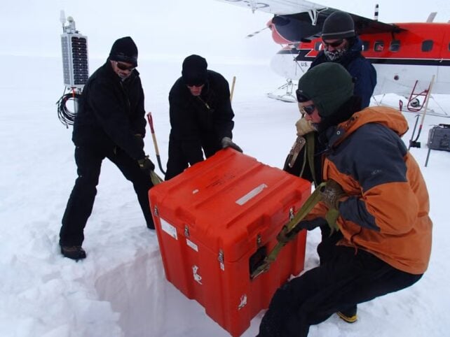 Researchers in cold-weather gear unload a large orange equipment case from a plane onto a snowy Antarctic field site.
