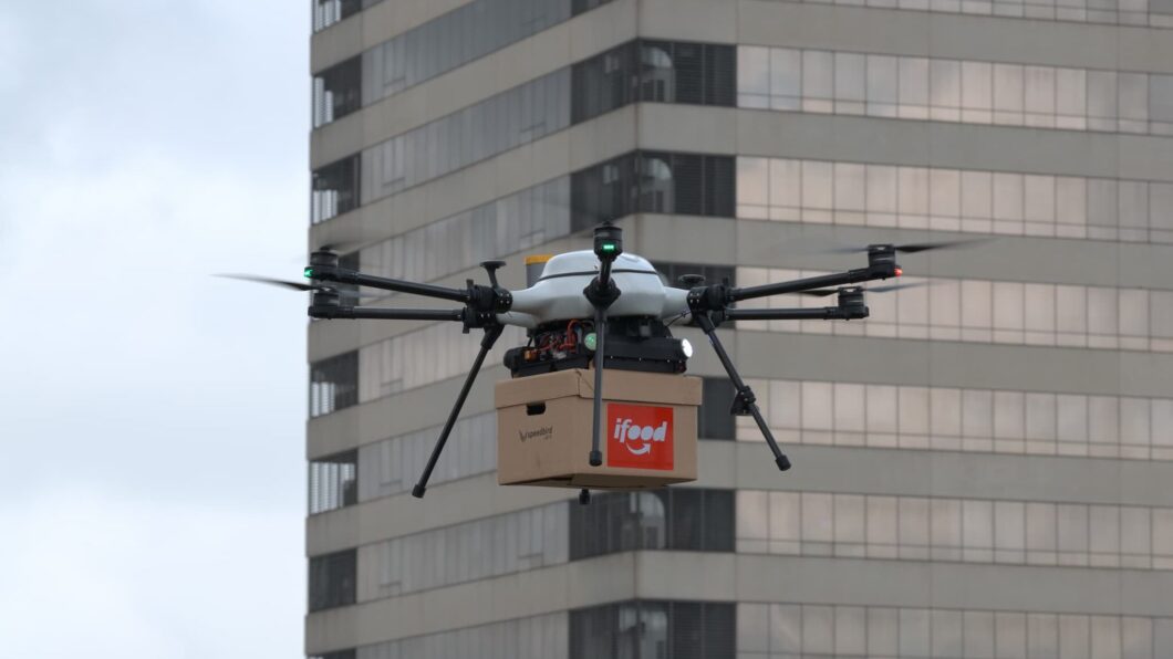 Foto em plano médio de um drone multi-rotor branco e preto voando de perfil, em um dia nublado. O drone carrega acoplada em sua base uma caixa quadrada de papelão pardo, contendo um adesivo retangular vermelho com o logotipo da marca "iFood" escrito em letras brancas. À esquerda do adesivo, há o logotipo estilizado e o nome da empresa "speedbird aero" gravados em preto na própria caixa. Ao fundo, vê-se a fachada de um edifício residencial ou comercial alto com janelas espelhadas.
