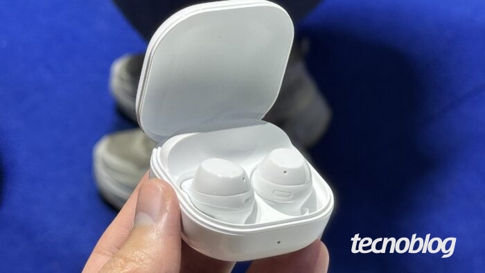 Uma mão segura um estojo de fones de ouvido sem fio de cor branca, que está aberto e revela dois fones também brancos. O logotipo "tecnoblog" está no canto inferior direito, em branco.