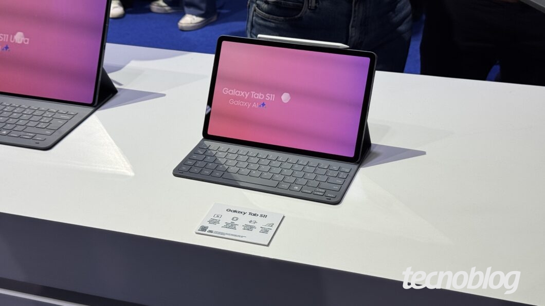 Dois tablets Galaxy Tab S11 estão em exposição em um balcão branco, um em primeiro plano e o outro no fundo. O tablet em primeiro plano está conectado a um teclado, com uma caneta Stylus magnética fixada na lateral. A tela exibe um fundo em gradiente de tons de roxo, rosa e vermelho, e mostra as palavras "Galaxy Tab S11" e "Galaxy AI" com um ícone de estrela. Um pequeno folheto de informações com as mesmas palavras está na frente do dispositivo. O logotipo "tecnoblog" está no canto inferior direito.