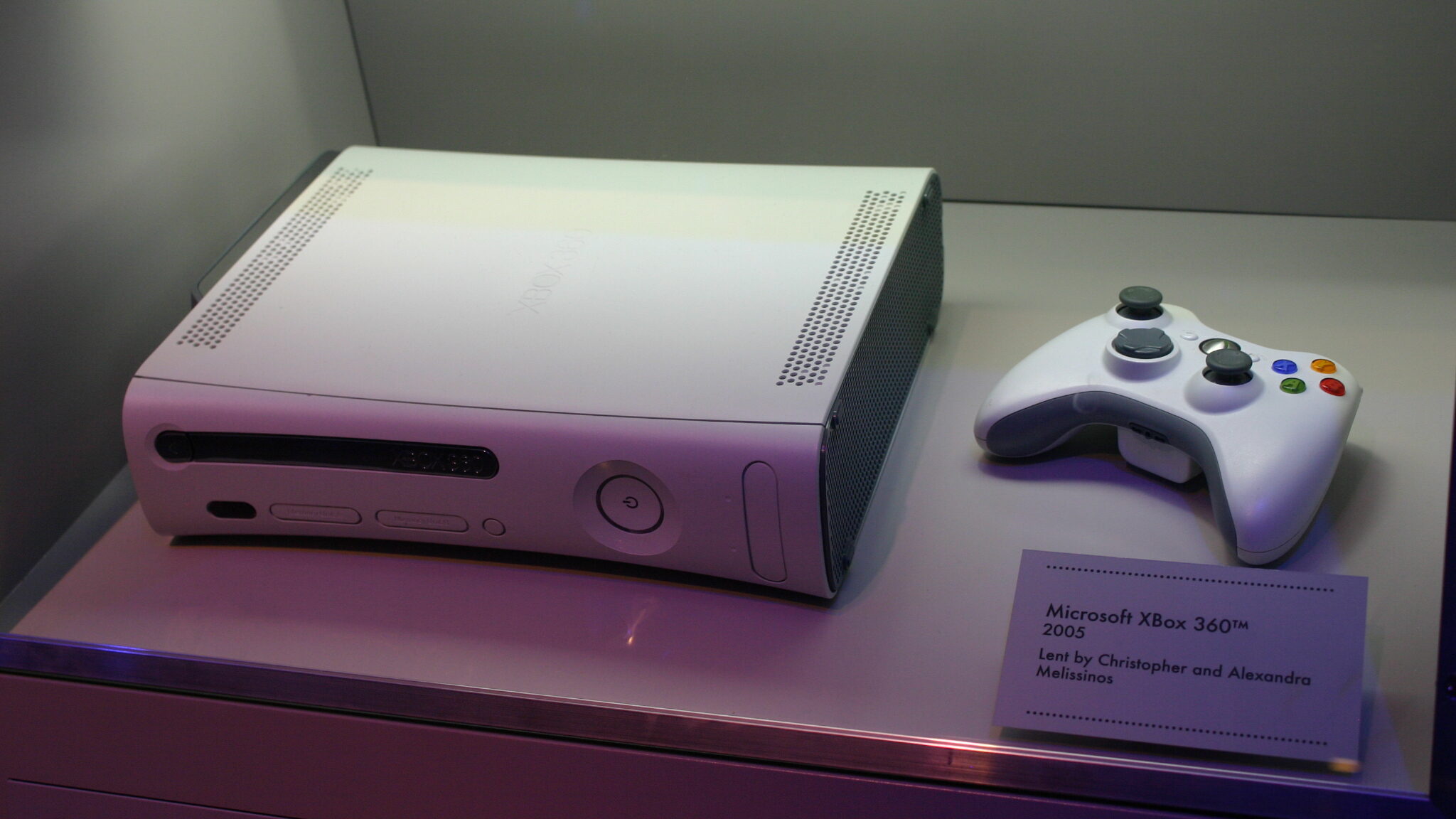 Console Microsoft Xbox 360 na cor branca, com controle e placa “Microsoft Xbox 360™ 2005”. Produto é exibido em uma feira de tecnologia.