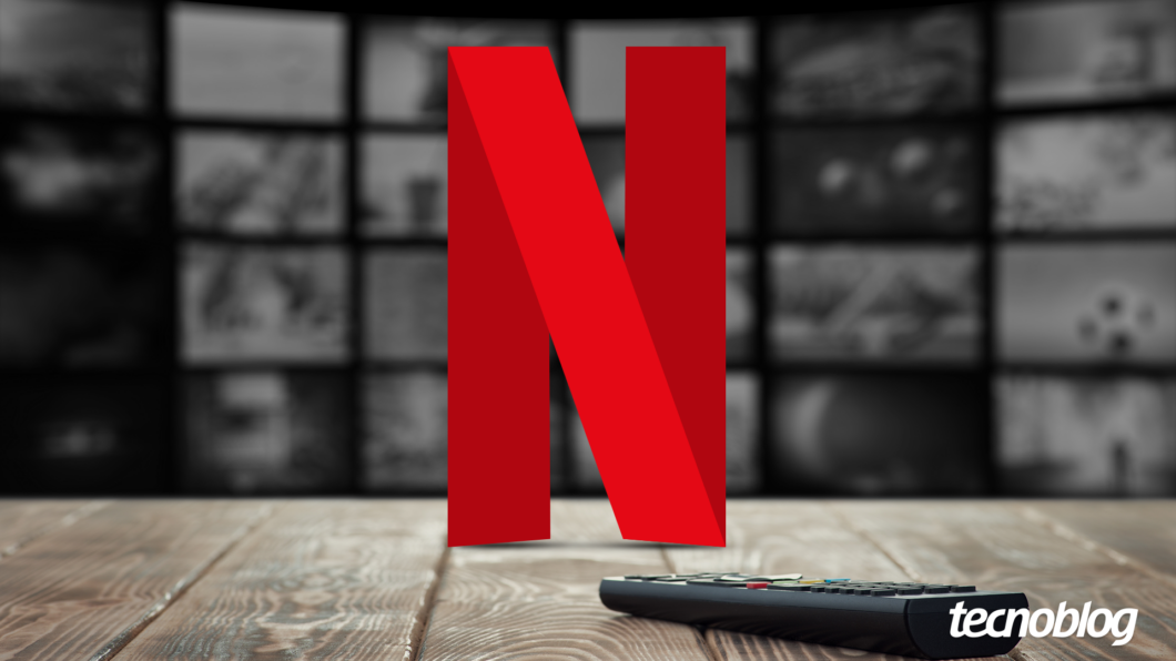Imagem mostra o logo vermelho estilizado da Netflix, um "N" grande com uma curva na parte inferior. Abaixo e ligeiramente à direita do logo, um controle remoto preto está parcialmente visível. Ao fundo, uma parede composta por várias telas de televisão desfocadas exibe diversas cenas de filmes e séries em tons de cinza. Na parte inferior direita, o logotipo do "Tecnoblog" é visível.