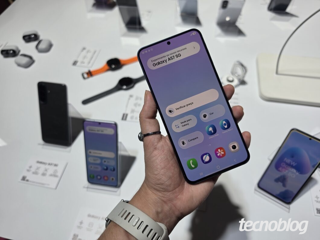 Pessoa segurando um celular Samsung Galaxy A57 5G exibindo a tela inicial, ao lado de outros modelos em uma bancada de exposição
