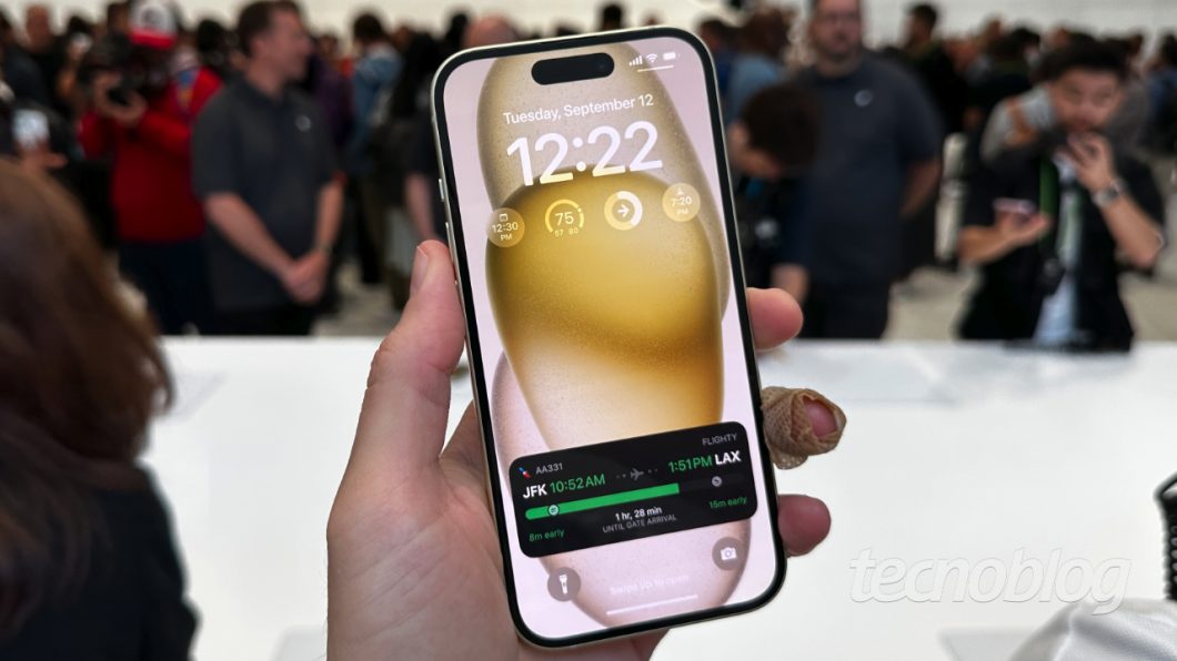 iPhone 15 sendo exibido na mão, com tela inicial e notificações em ambiente de evento