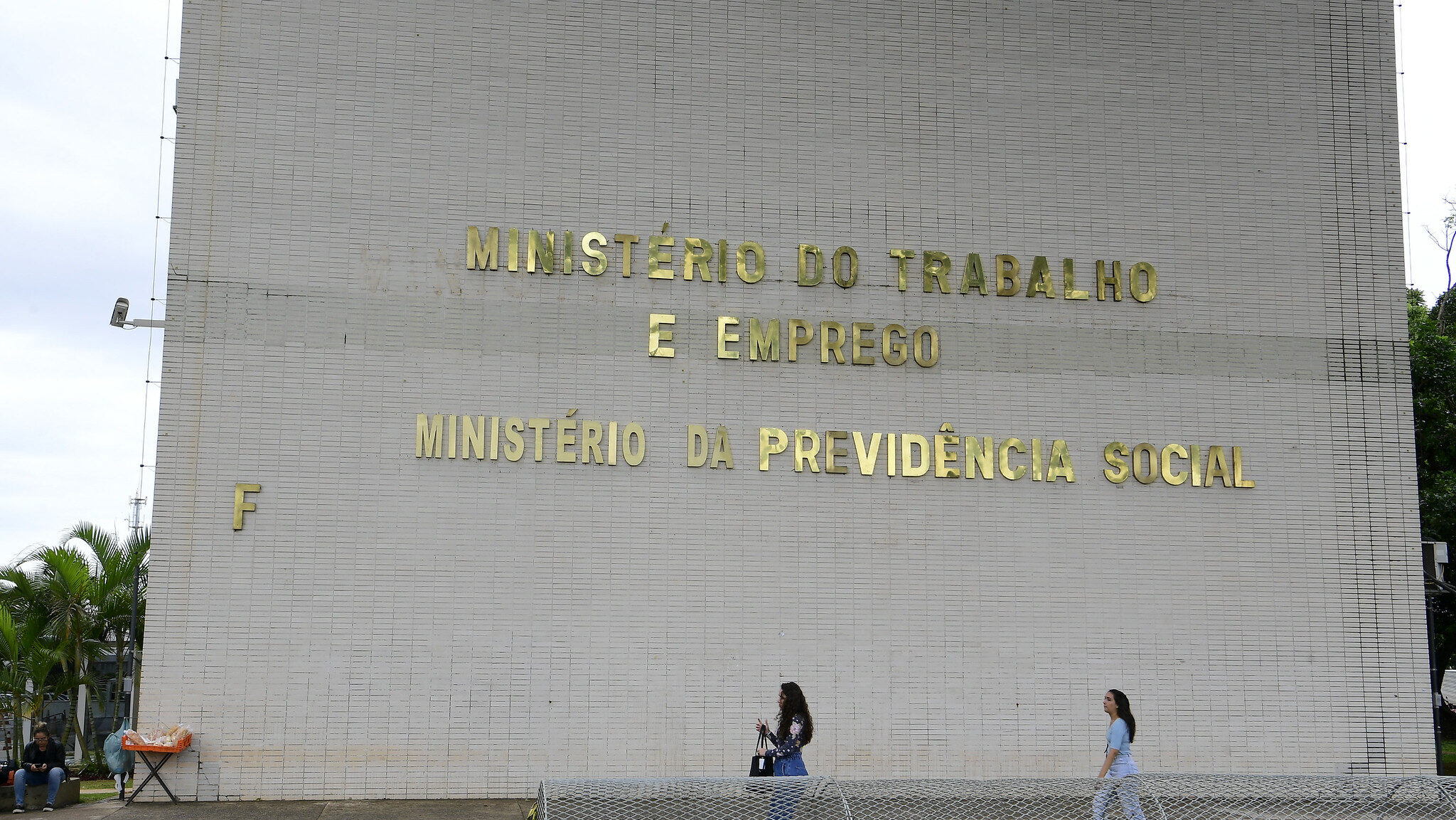 Fotografia da fachada lateral de um prédio público com paredes de pastilhas claras. No centro, letras metálicas douradas formam o texto "MINISTÉRIO DO TRABALHO E EMPREGO" e, logo abaixo, "MINISTÉRIO DA PREVIDÊNCIA SOCIAL". À esquerda, a letra "F" dourada sobressai na parede. Na calçada em frente, duas pessoas caminham paralelas ao prédio, atrás de uma barreira cilíndrica de concreto e metal. No canto esquerdo inferior, outra pessoa está sentada ao lado de uma mesa dobrável.