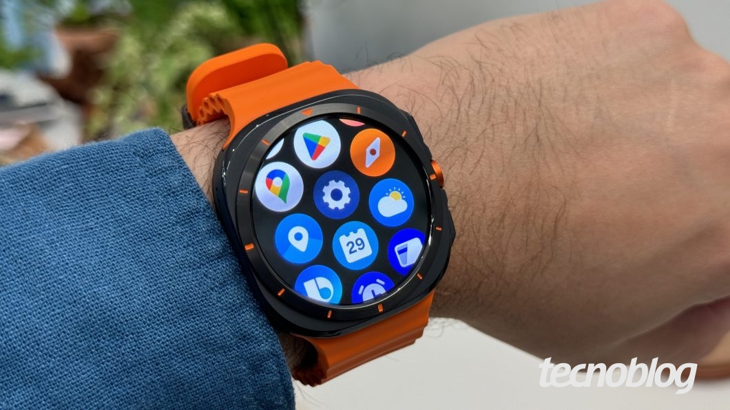 Mostrando o painel de aplicativos de um smartwatch laranja no pulso, com ícones de navegação, clima e mensagens