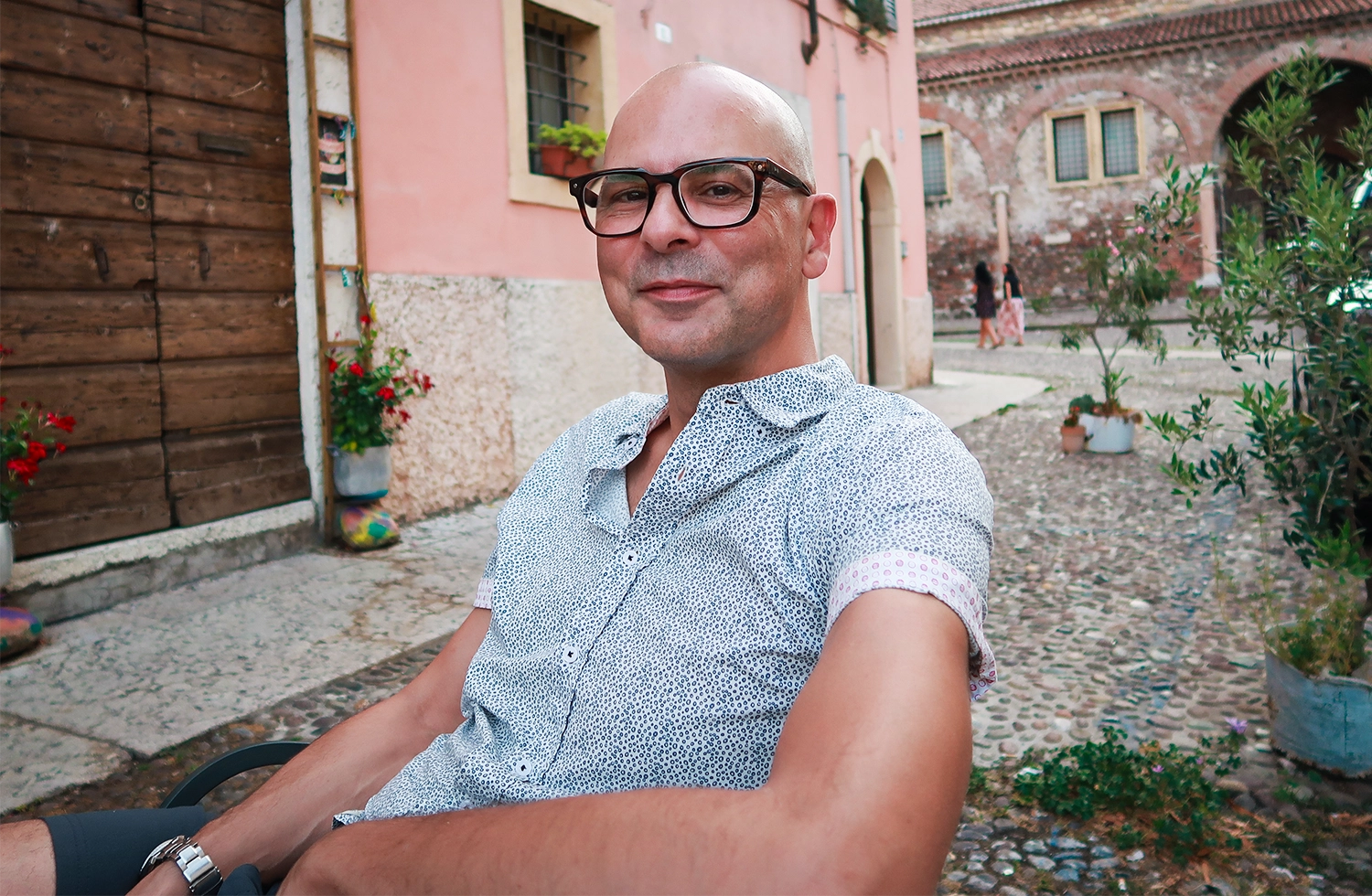 A man wearing glasses and a light blue shirt sits in front of a pale pink building.