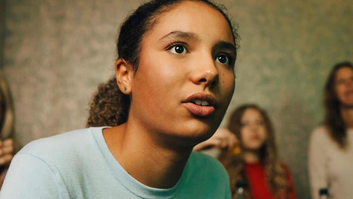 Close-up of a teenage girl leaning forward with a focused expression while watching something off-camera, with other people blurred in the background.