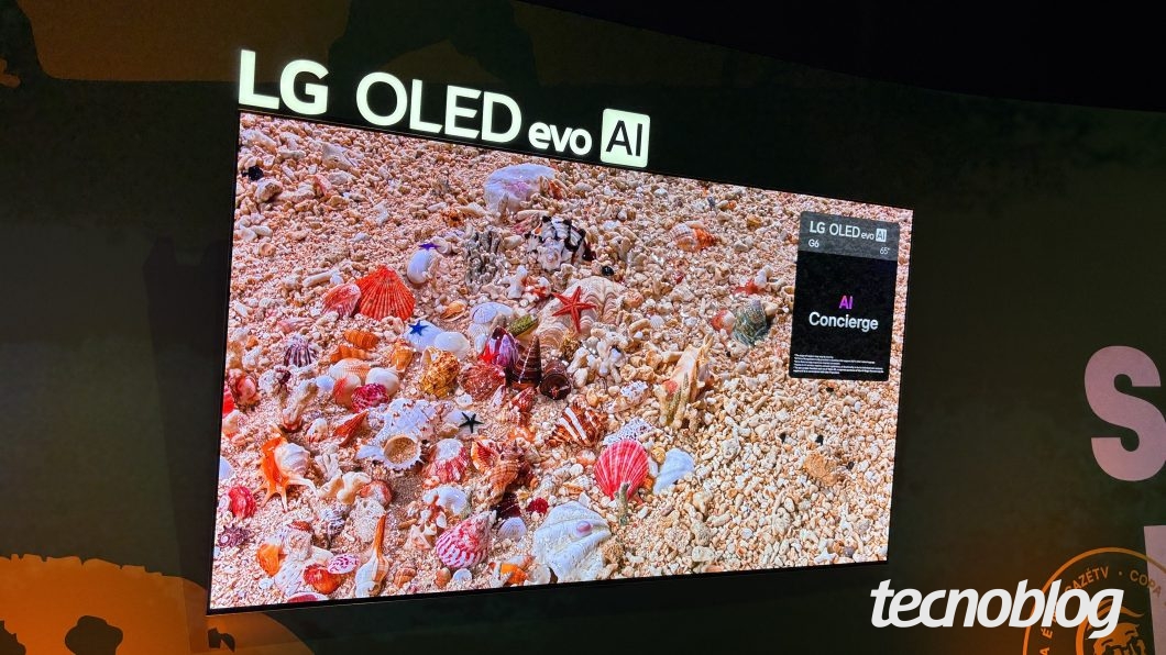 Smart TV da LG instalada em parede escura durante evento. Acima da tela, há uma placa iluminada com o texto "LG OLED evo AI". No display, é exibida uma imagem em alta definição repleta de conchas e estrelas-do-mar coloridas sobre areia clara. No canto superior direito da tela, um card preto exibe os textos "LG OLED evo AI", "G6", "65"", "AI Concierge" e pequenas linhas explicativas. No canto inferior direito da cena, nota-se parte de um selo circular.