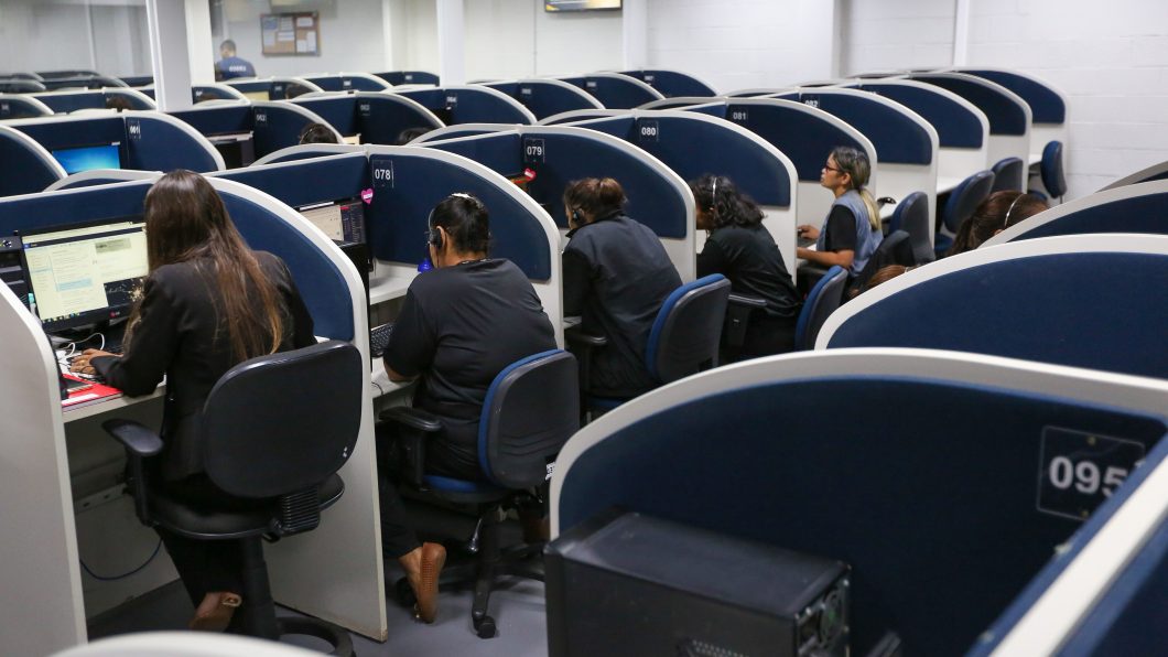 Imagem mostra uma central de atendimento, com pessoas sentadas, de costas para a câmera, interagindo com computadores em cabines