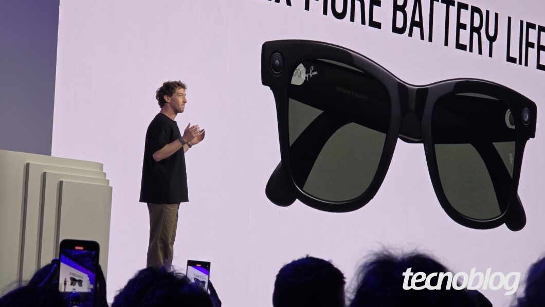 Mark Zuckerberg aparece de perfil no lado esquerdo de um palco iluminado, vestindo uma camiseta preta e calças cáqui, gesticulando enquanto fala. Na parte inferior direita, o logotipo do "tecnoblog" é visível.