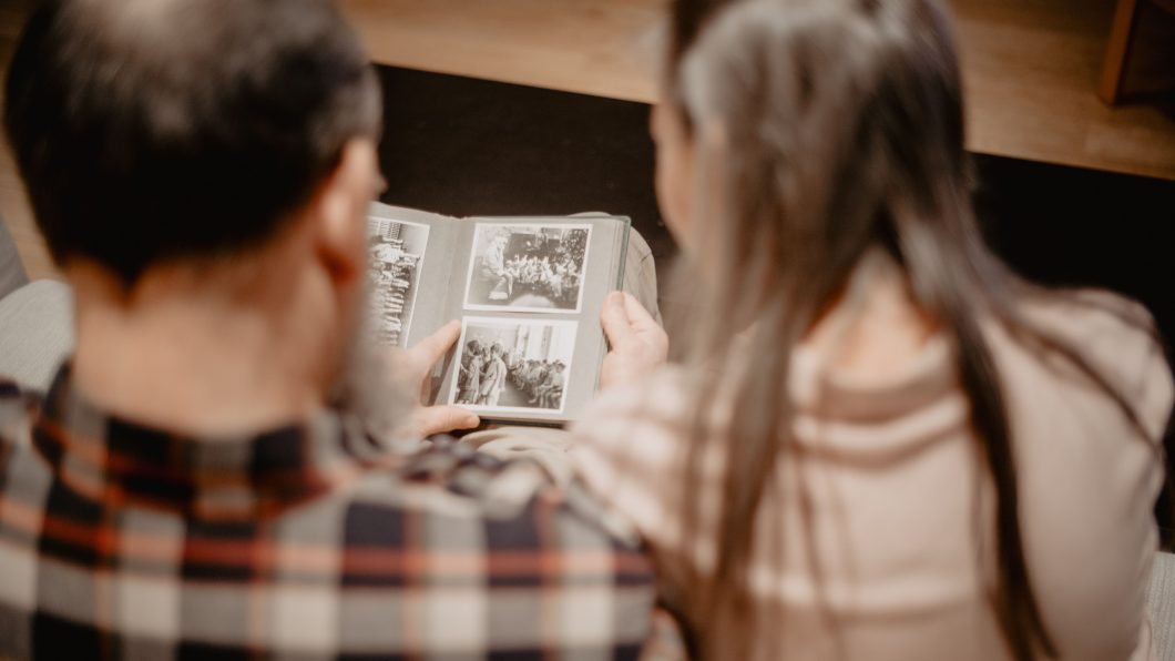 Fotografia em plano médio com iluminação acolhedora, tirada do ponto de vista posterior de um casal sentado lado a lado. Um homem vestindo camisa xadrez em tons de marrom e preto aponta o indicador para a página de um álbum de fotos impresso pequeno, enquanto a mulher à direita, de cabelos castanhos longos e blusa bege, observa atentamente as fotografias antigas.