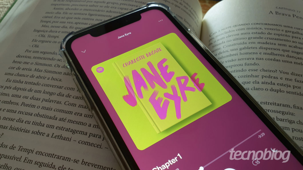 Fotografia em plano detalhe mostra um smartphone com capinha transparente repousando sobre as páginas abertas de um livro impresso; a tela exibe o aplicativo Spotify executando o audiobook do romance Jane Eyre, de Charlotte Brontë. Na parte inferior direita, aparece a marca d'água "tecnoblog"